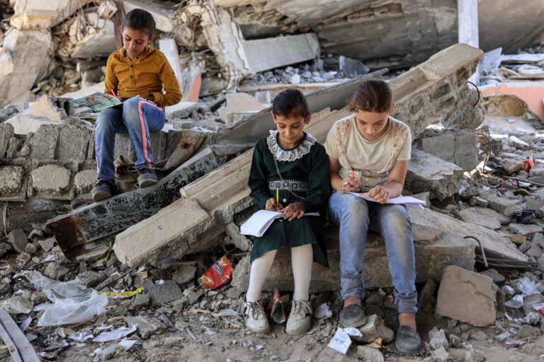 113138901-files-children-write-in-notebooks-by-the-rubble-of-destroyed-buildings-near-a-tent-being-u.jpeg